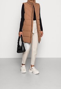 Kvinna iklädd en brun vadderad ärmlös kappa, svart långärmad topp, vita byxor, vita sneakers med leoparddetaljer, som håller en svart rynkad handväska.