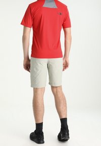 Camiseta corta roja que absorbe la humedad con panel trasero gris, combinada con pantalones cortos beige claro y zapatillas deportivas negras, vista desde la parte trasera.