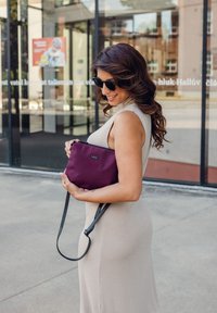 Fialová nylonová kabelka s černým nastavitelným popruhem, která se vyznačuje minimalistickým designem a malým logem na přední straně, držená osobou.