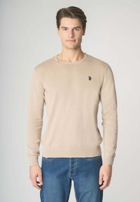 Maglione beige lavorato a maglia con scollo rotondo, maniche lunghe, polsini a costine e un emblema blu sul petto, indossato con jeans blu.