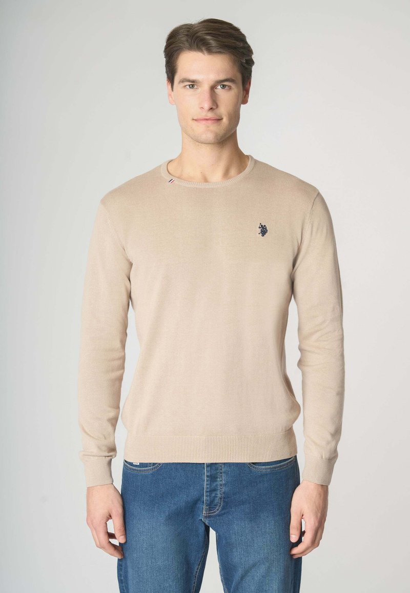 Maglione beige lavorato a maglia con scollo rotondo, maniche lunghe, polsini a costine e un emblema blu sul petto, indossato con jeans blu.