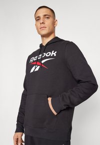 Crna dukserica s kapuljačom od mekog materijala, koja ima veliki bijeli i crveni Reebok logo, kengurú džep i rebraste manšete.