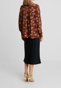 Chemise à manches longues bordeaux avec motif floral jaune et bleu foncé. Associée à une jupe mi-longue noire et des sandales à talons dorés.