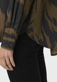 Camicia con un motivo astratto marrone e nero, dalla texture liscia, con maniche lunghe e polsi abbottonati. Abbinata a pantaloni neri con tasca.