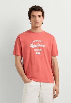 Junger Mann mit lockigem Haar, der ein korallenrotes Pepe Jeans T-Shirt mit weißem Aufdruck und cremefarbene Hose trägt, steht mit einer Hand in der Tasche.