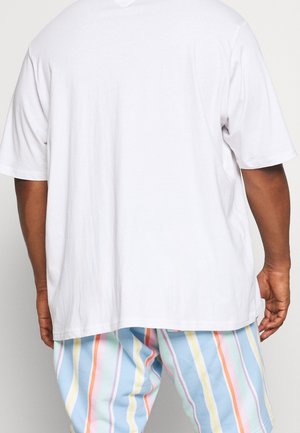 Person iført en løs hvid kortærmet skjorte og bukser med pastelstriber, vendt væk med hænderne afslappet ved siden.