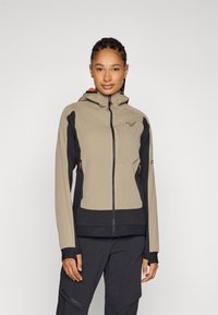 Beige en zwarte softshell jas met een capuchon, ritszakken en elastische manchetten. Heeft een logo op de borst en gestructureerde accenten.