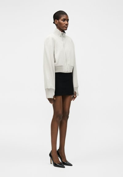 Γυναίκα όρθια με το προφίλ, φοράει ένα ανοιχτό γκρι ζιπ-παντελόνι, μαύρη μίνι φούστα, μαύρες γόβες και μικρά σκουλαρίκια πάνω σε λευκό φόντο.