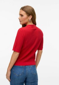 Röd stickad polo med korta ärmar, med ett strukturerat mönster. Bärs med blå jeans och visas från baksidan.