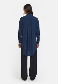 Chemise à manches longues bleu marine avec un ourlet incurvé et un pli au dos, fabriquée en tissu léger. Associée à un pantalon noir large.