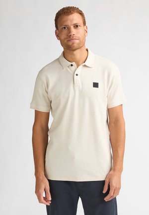 Polo beige en tissu texturé, patte de boutonnage à trois boutons et petit logo carré noir sur la poitrine. Manches courtes et coupe décontractée.