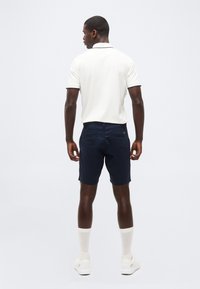 Άνδρας που στέκεται με την πλάτη προς τα εμπρός, φοράει λευκό πόλο με κοντά μανίκια, σκούρα μπλε σορτς, λευκές κάλτσες μέχρι τους αστραγάλους και λευκά αθλητικά παπούτσια.