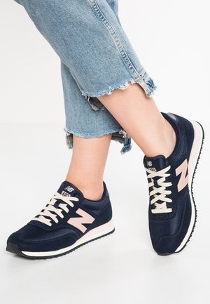 Piernas con vaqueros cortos azules y zapatillas New Balance azul marino con cordones blancos y logo rosa claro, de pie sobre una superficie blanca.