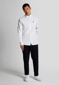 Camisa blanca de manga larga con botones y un bolsillo en el pecho, adornada con un logo de corazón amarillo, combinada con pantalones negros y zapatillas blancas.