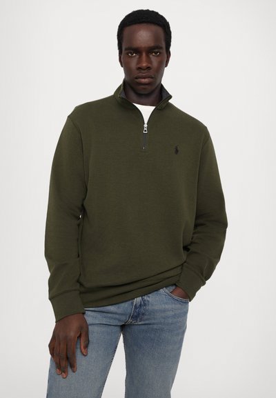 Grøn kvart-lynlås sweatshirt med lange ærmer, præget af et logo på brystet. Båret over en hvid t-shirt, parret med blå jeans.