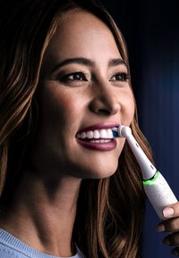 Brosse à dents électrique au corps blanc avec un design moucheté, des poils bleus et une lumière LED verte à la base, tenue près d'une bouche souriante.