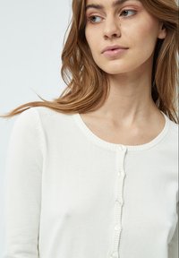 Giovane donna con capelli castano chiaro che indossa un cardigan bianco abbottonato, che guarda leggermente di lato su uno sfondo semplice.