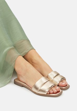 Fémes arany slip-on szandál széles, párnázott felsőrész pántokkal és lapos, téglalap alakú talppal. Átlátszó zöld ruhával viselve.