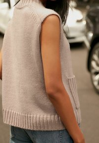 Jasnoróżowy bezrękawnik z dzianiny z prążkowanym dołem i boczną kieszenią, z wysokim kołnierzem i miękką teksturą. Noszony na niebieskich jeansach.