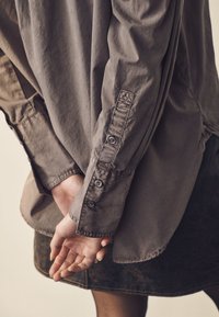 Gray button-up shirt with long sleeves, featuring a loose fit, soft texture, and subtle stitching details, paired with a dark skirt.