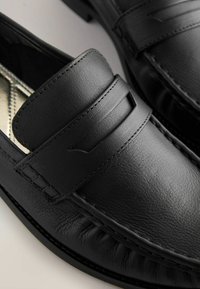 Black leather loafers feature a stitched design, a horizontal strap across the upper, and a smooth texture with a slight sheen.