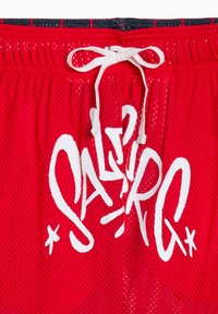 Shorts en maille rouge avec taille à cordon de serrage, arborant un grand motif graphique blanc avec "SABIR" écrit sur le devant.