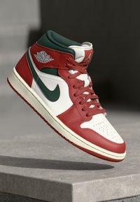 Crvene, bijele i tamnozelene Air Jordan 1 tenisice lebde iznad betonske površine s vidljivim Nike swoosh i Air Jordan logotipima.