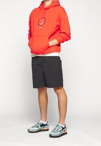 Röd bomulls-hoodie med framficka och svart grafiskt tryck. Kombinerad med svarta shorts och sneakers i turkos, svart och orange.