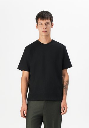 Black short-sleeve crewneck t-shirt made of cotton, featuring a relaxed fit and smooth texture, worn with olive green pants, minimalistic design.