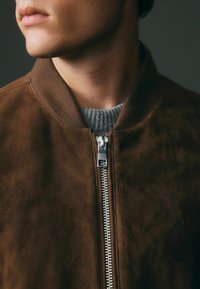 Chaqueta bomber de ante marrón con cierre de cremallera, cuello acanalado y un suéter gris de punto texturizado debajo. Los acentos incluyen una cremallera plateada.