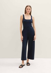 Mono azul marino con escote cuadrado, diseño de pierna ancha y textura suave, combinado con sandalias planas negras sobre un fondo claro.