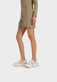 Sneaker atletici bianchi con accenti grigio chiaro, dotati di una tomaia texturizzata, suola liscia e lacci. Indossati con un vestito aderente verde oliva.