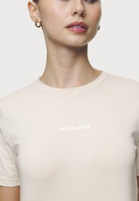 Beige bavlněné tričko s kulatým výstřihem a krátkým rukávem. Na přední straně se nachází bílý nápis "PEGADOR" v tučném písmu bez patky.