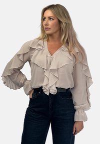 Jasnoróżowa bluzka z falbankami, dekoltem w kształcie litery V oraz długimi bufkami. Połączona z ciemnymi dżinsami, które podkreślają warstwową teksturę.