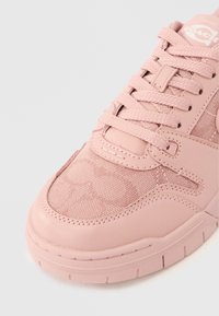 Ροζ αθλητικό παπούτσι Coach με υφασμάτινη υφή και δερμάτινες λεπτομέρειες, δεμένα κορδόνια και ορατό λογότυπο στη γλώσσα, απεικονίζεται από κοντινή γωνία.