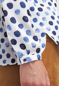 Blusa a maniche lunghe bianca con motivo a pois blu e navy. Presenta un polsino con bottone e uno spacco laterale. Tessuto morbido e testurizzato.