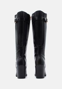 Bottes noires montant jusqu'au genou en cuir lisse, présentant des détails de couture verticale, un accent de boucle près du haut et un talon épais.