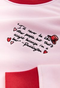 Pink fabric with embroidered quote about Paddington bear, red hearts, and a red hat, with red ribbed trim visible at edges.