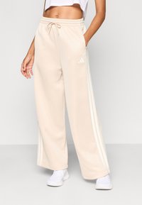 Bež široke trenirke iz mehkih materialov, z belimi stranskimi črtami in elastičnim pasom z vrvico za zavezovanje.