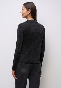 Musta pitkähihaine paita, jossa on hillitty kiilto ja kaulus, yhdistettynä harmaisiin housuihin, katsottuna takaa vaaleaa taustaa vasten.