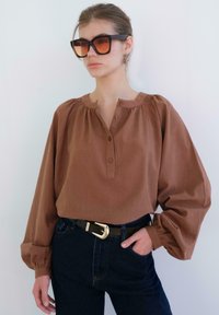 Žena v nadměrném hnědém topu s balónovými rukávy, tmavých džínách, černém opasku se zlatou sponou a velkých hranatých slunečních brýlích, stojí proti bílé zdi.
