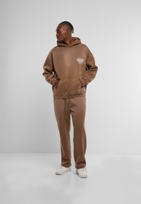 Brauner Pullover und passende Sweatpants aus weichem Stoff. Verfügt über eine Fronttasche und einen Grafikdruck auf der Brust. Weiße Sneaker.