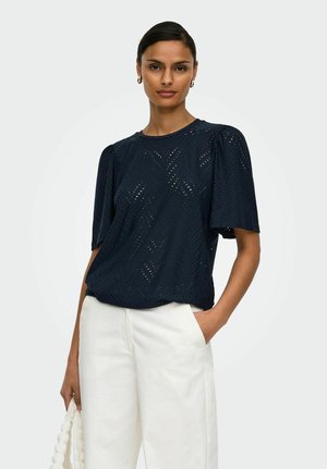 Kvinde iført en sort perforeret bluse med korte ærmer og chevronmønstre samt hvide bukser med høj talje, der holder en hvid tekstureret taske.