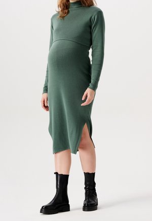 Těhotná žena nosící zelené pletené midi šaty s dlouhým rukávem a bočním rozparkem, černé kotníkové boty, stojící před jednobarevným pozadím.