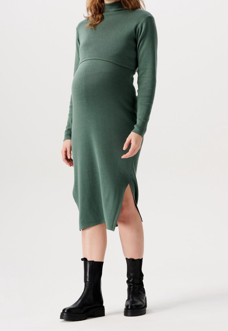 Femme enceinte portant une robe midi en maille verte à manches longues avec une fente latérale et des bottines noires, debout devant un fond uni.