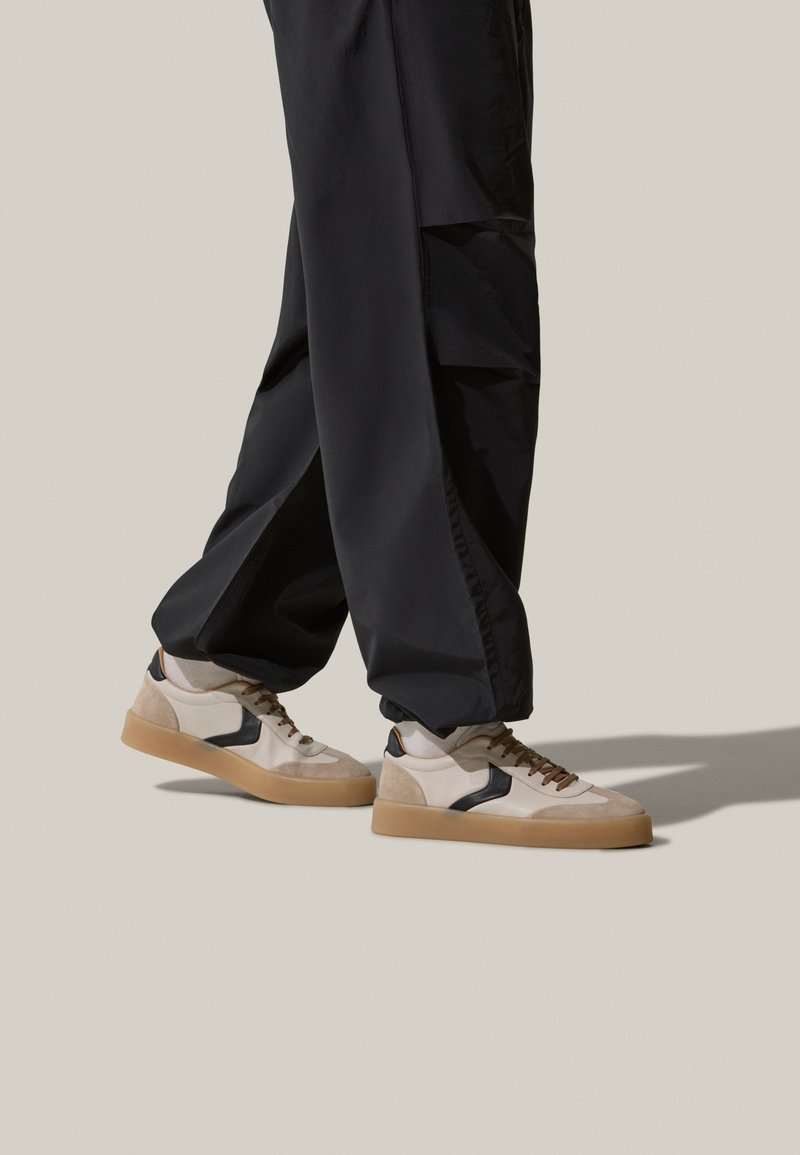 Sneakers beige e neri realizzati in materiali scamosciati e in pelle. Presentano una punta rotonda, suola in gomma e lacci marroni. Indossati con pantaloni neri.