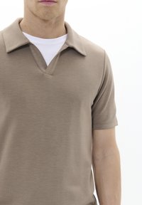 Krátký rukáv hnědý polo triko s výstřihem do V, měkkou texturou a vrstvou bílého trička pod ním. Jednoduchý a minimalistický design.