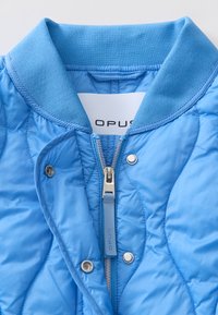 Veste bomber matelassée bleu clair avec col côtelé, fermeture éclair et étiquette de marque à l'intérieur. Texture lisse avec finition brillante.