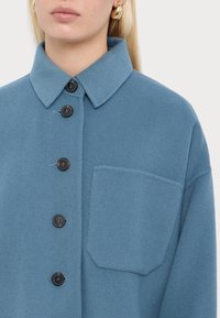 Giacca in pile blu con colletto, chiusura frontale a bottoni, dettagli di tasche finte e bottoni neri. Texture morbida e vestibilità rilassata.