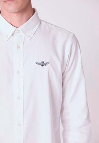 Camicia bianca con bottoni realizzata in cotone, dal texture morbido. Presenta un piccolo emblema azzurro scuro con ali all'altezza del lato sinistro del petto.